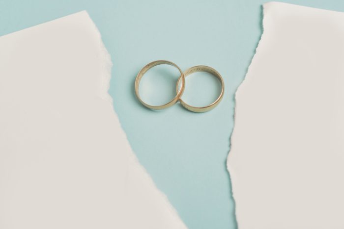 This image features two interlinked gold wedding rings resting on a soft blue background, framed by two pieces of torn white paper. The torn paper powerfully symbolises the breakdown of a marriage and the separation of a couple, while the enduring connection of the rings highlights the legal and financial ties that remain even after a relationship ends.