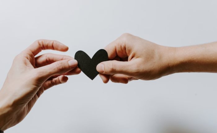Two hands holding a small black paper heart against a plain white background, symbolising the shared assets and legal considerations of an engaged couple.